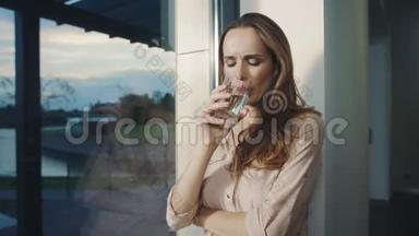 放松的女人在全景窗口附近喝水。 口渴的女士喝水